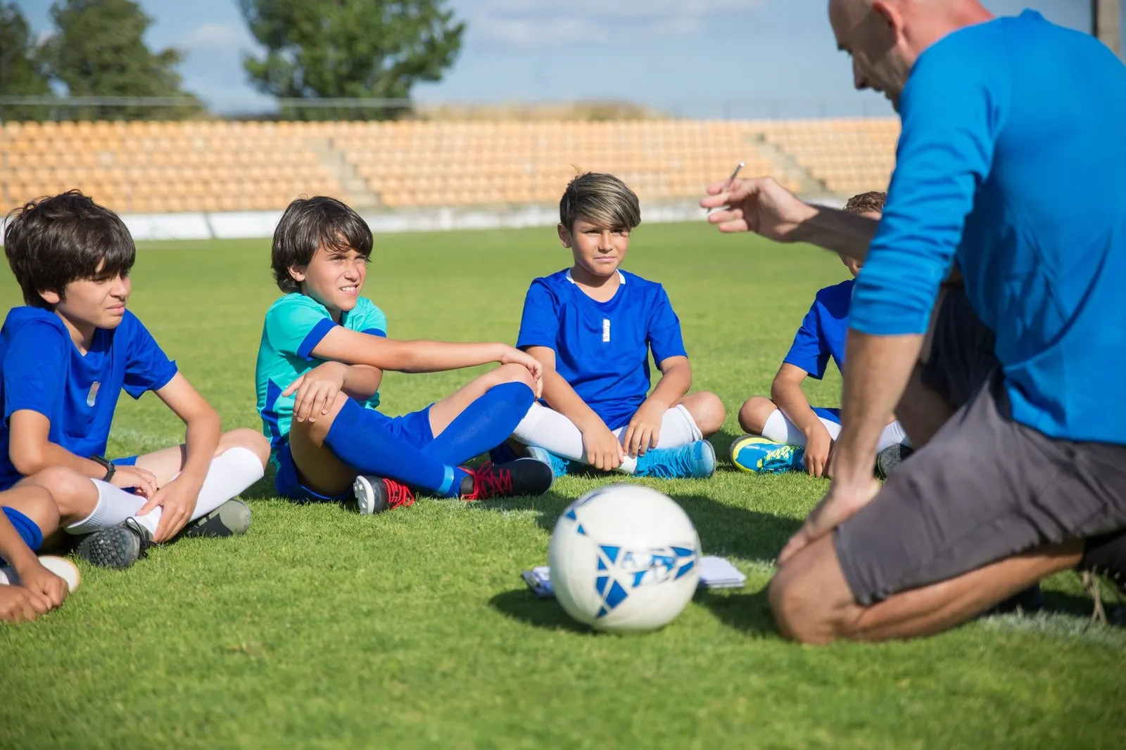 Băieți care își ascultă antrenorul de fotbal pe un teren însorit.